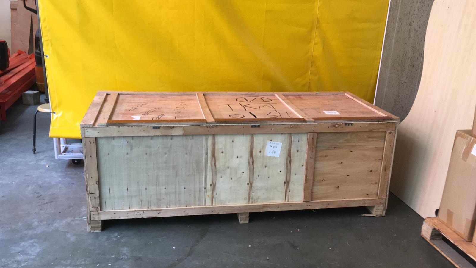 Large wooden crate with handwritten label sitting on a pallet across a concrete floor.