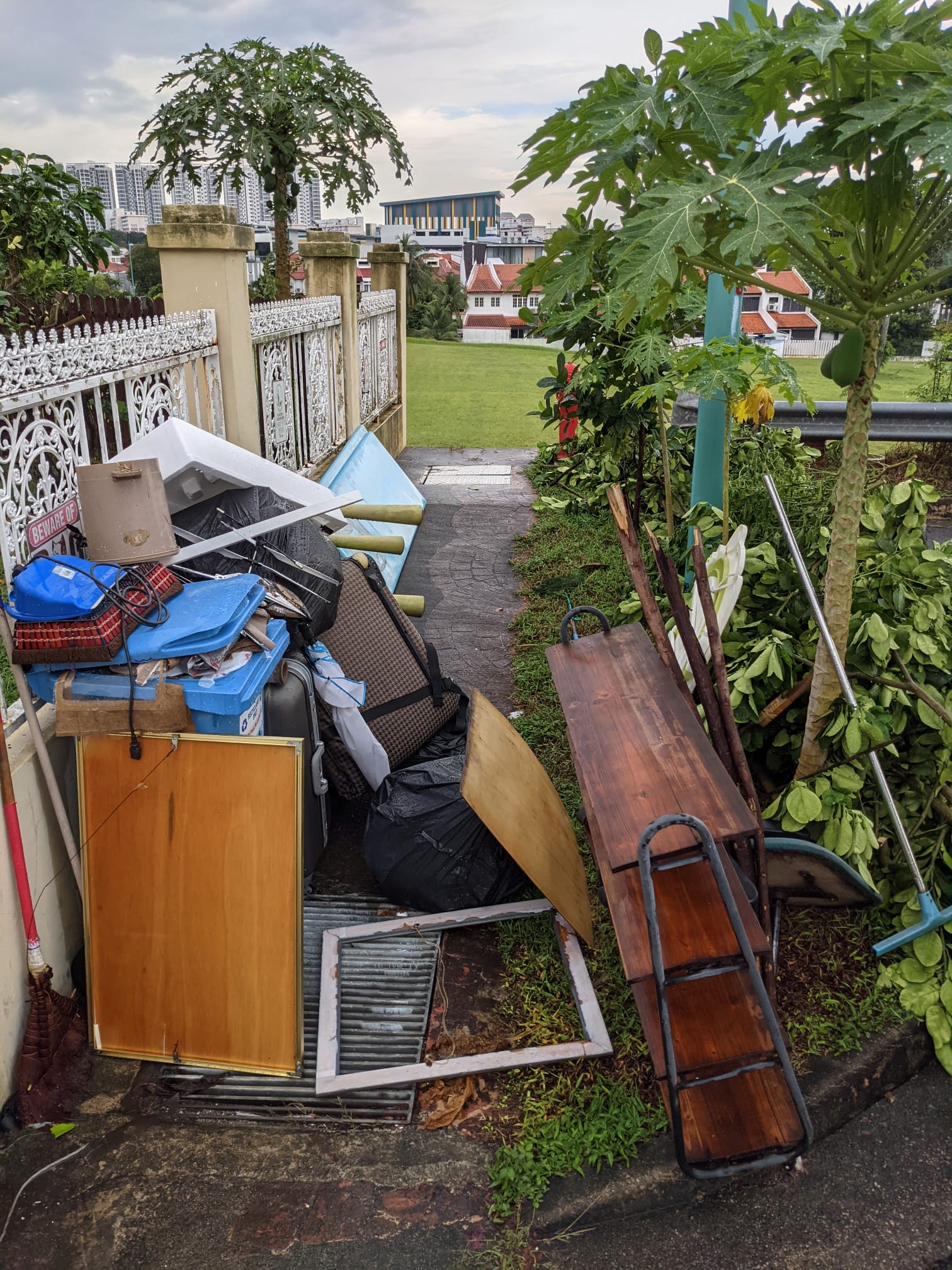 Pile of household furniture, plastic bags, and wooden debris stacked against a residential fence with trees in the background.