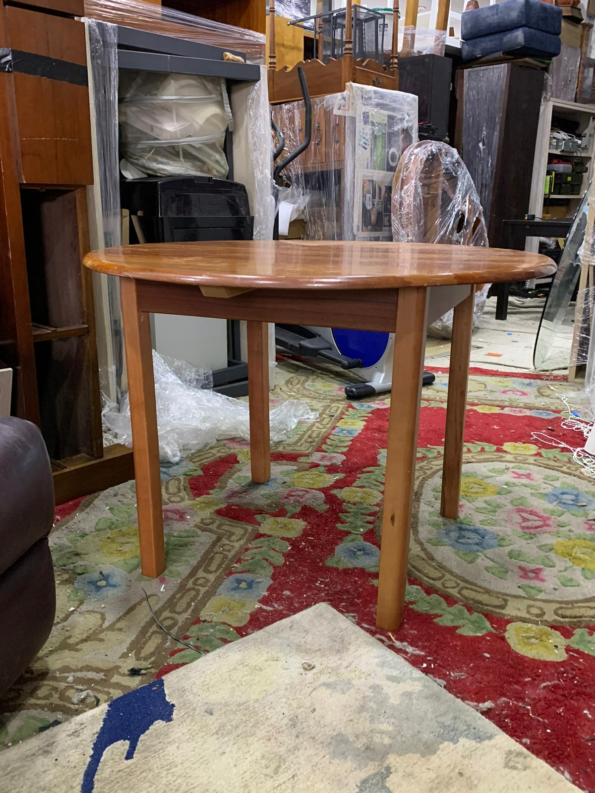 Wooden dining table with four legs positioned on a patterned rug, surrounded by plastic-wrapped furniture and household items.