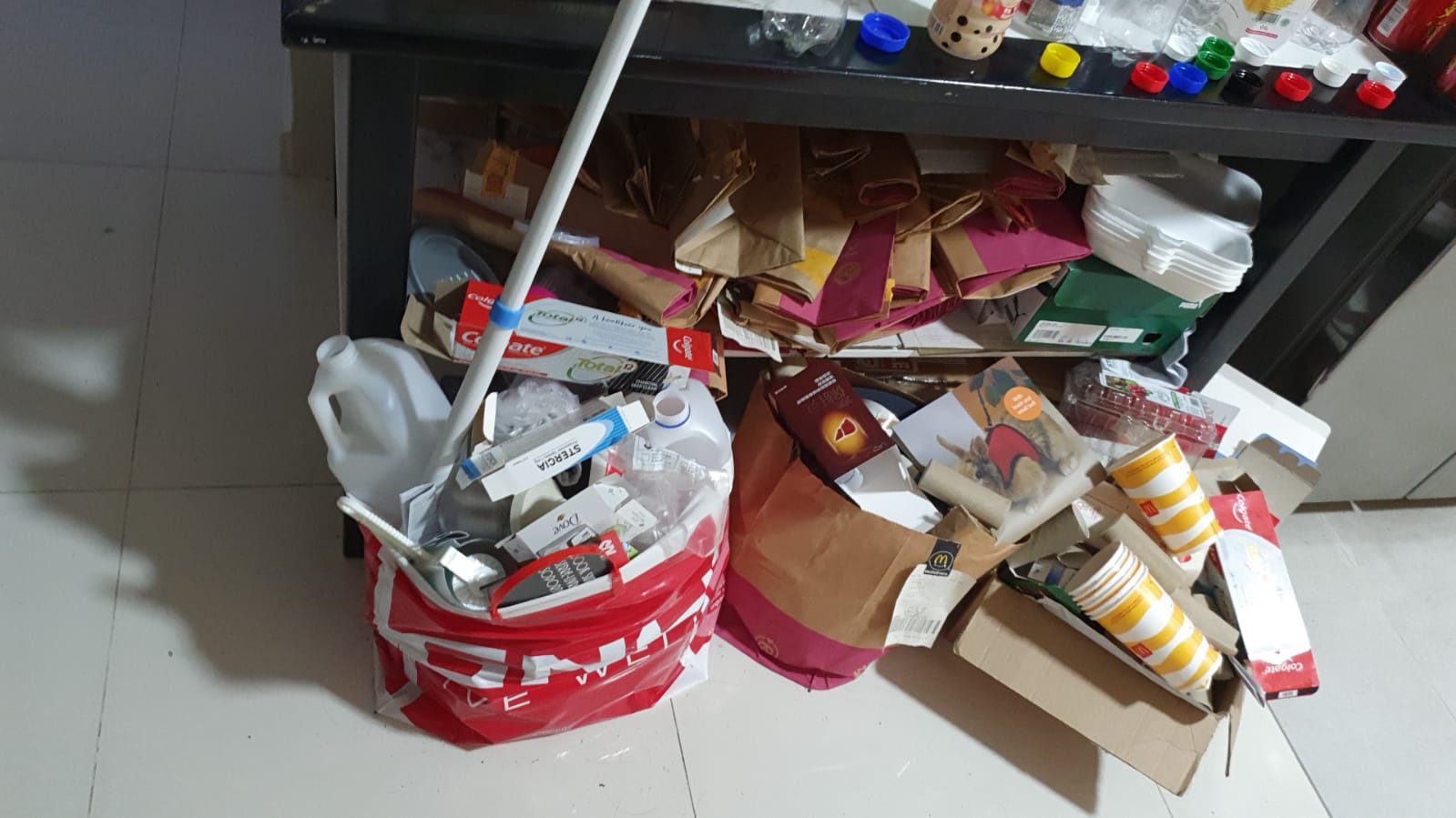 Plastic bottles, paper bags, and cardboard boxes accumulated on a residential floor beneath a shelf.