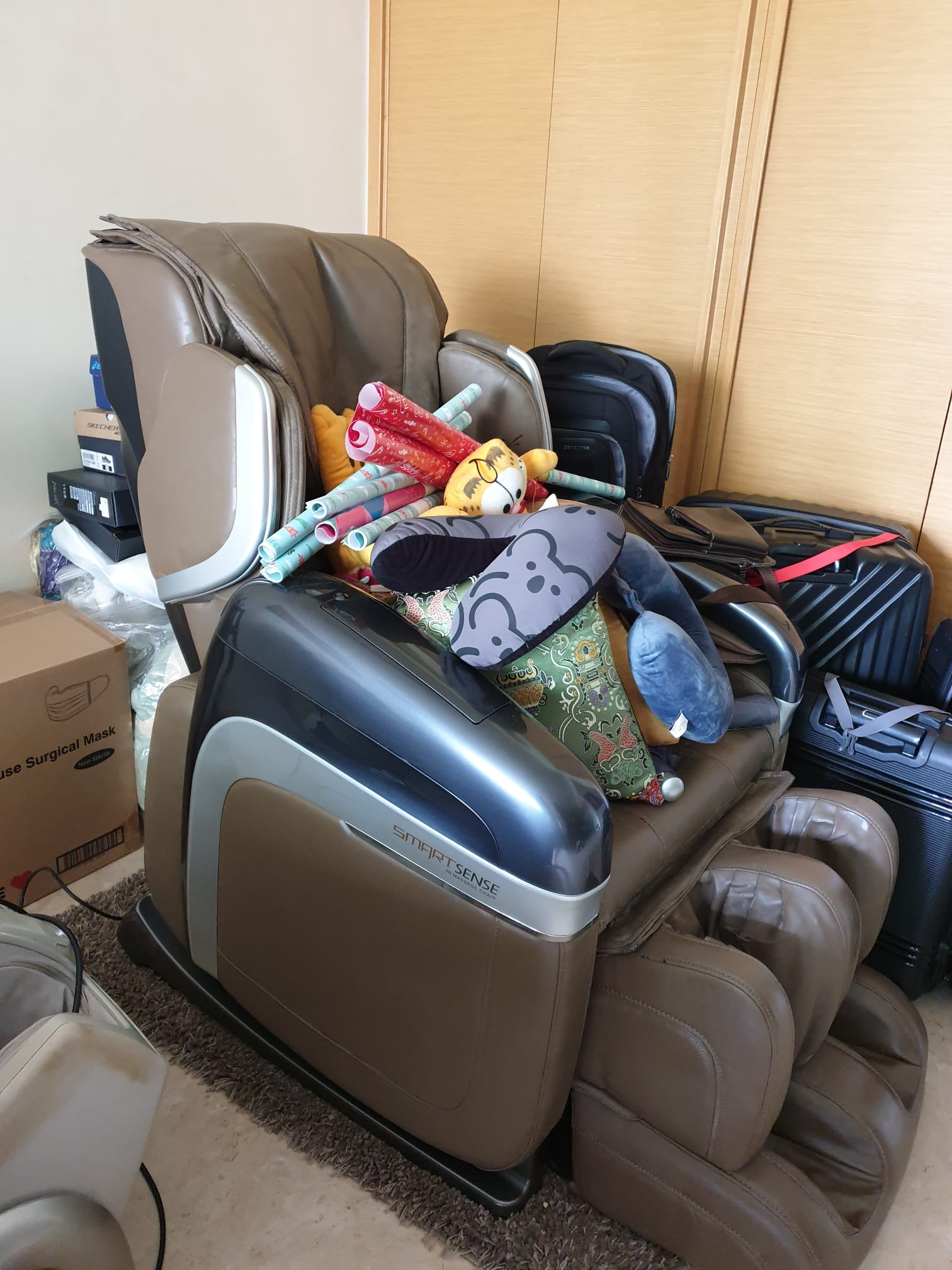 Massage chair surrounded by stacked pillows, rolled wrapping paper, and luggage pieces in a residential room.