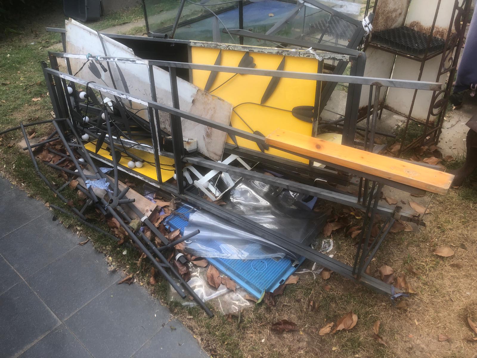 Heap of metal frames and structural debris stacked on grass beside a concrete path, with yellow warning signage nearby.