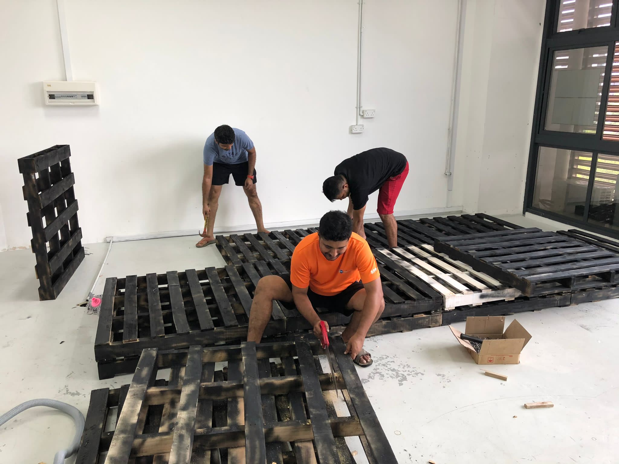 Two workers assembling a platform from dark wooden pallets in a large, empty indoor space.