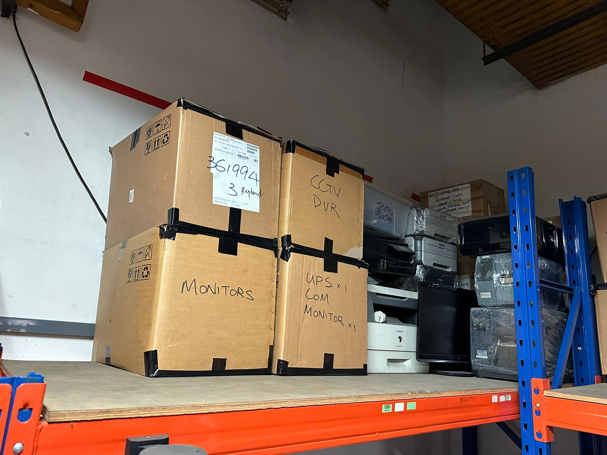 Stacked cardboard boxes marked MONITORS and CCTV DVR with printers and other electronics in a storage area.